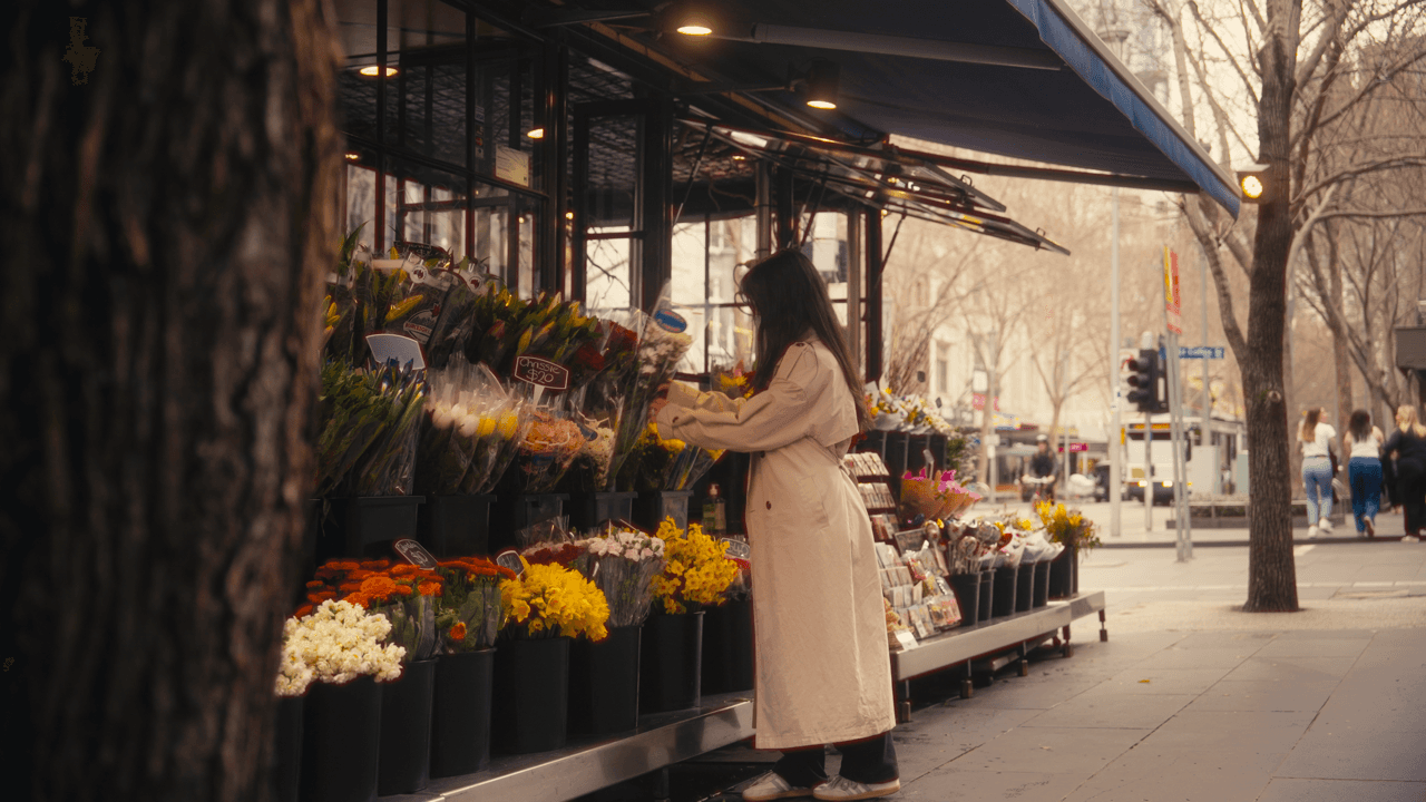 A woman picking flowers at an outdoor market stall, surrounded by a variety of vibrant flowers and greenery