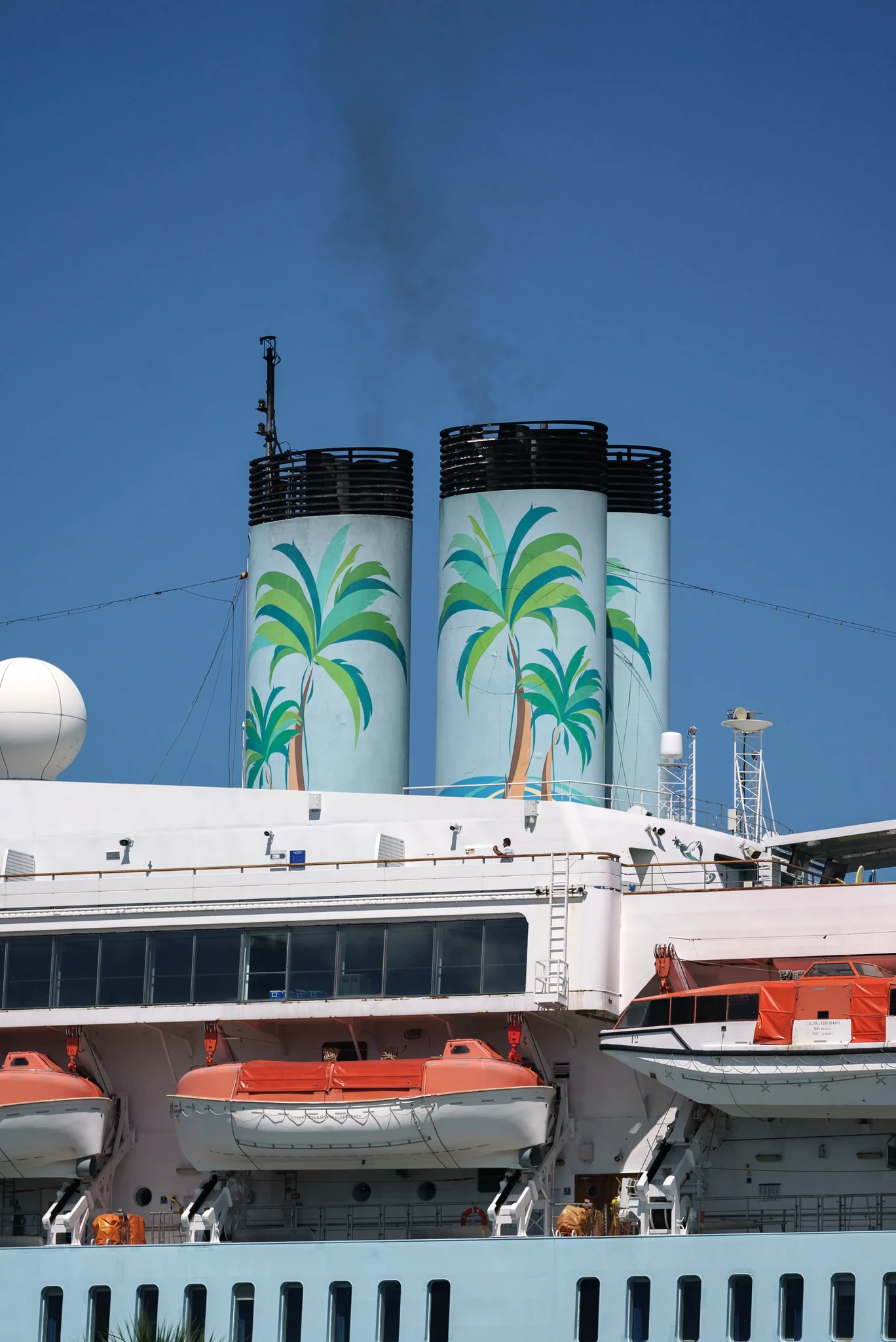 A cruise ship chimney with large palm tree illustrations on a sunny day. Smoke is visible coming from the top. Lifeboats are lined up below, painted in orange and white. The sky is clear and blue.