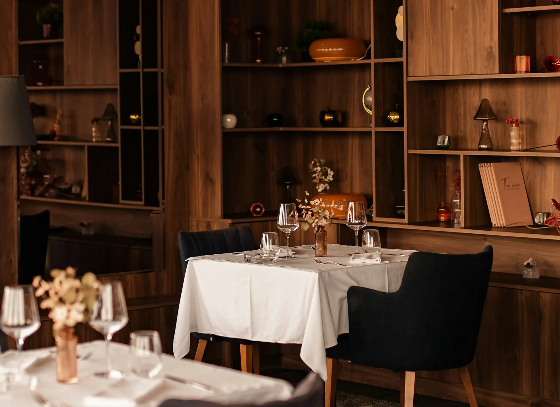 Modern restaurant interior with wooden shelving, elegant table setting, wine glasses, and warm ambient lighting creating an intimate dining atmosphere.