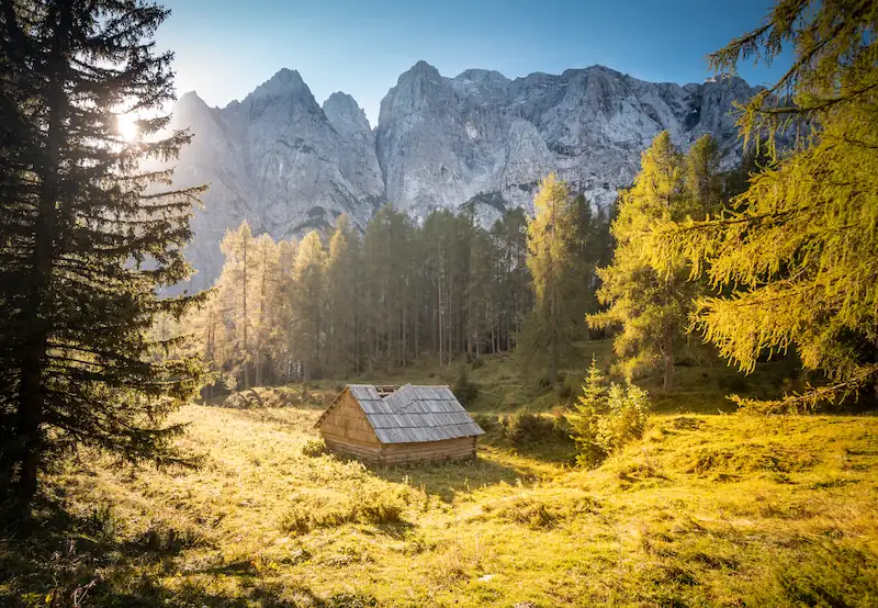 Sunlight hitting a small mountain hut in a golden meadow, surrounded by epic mountains of Vršič pass, Slovenia