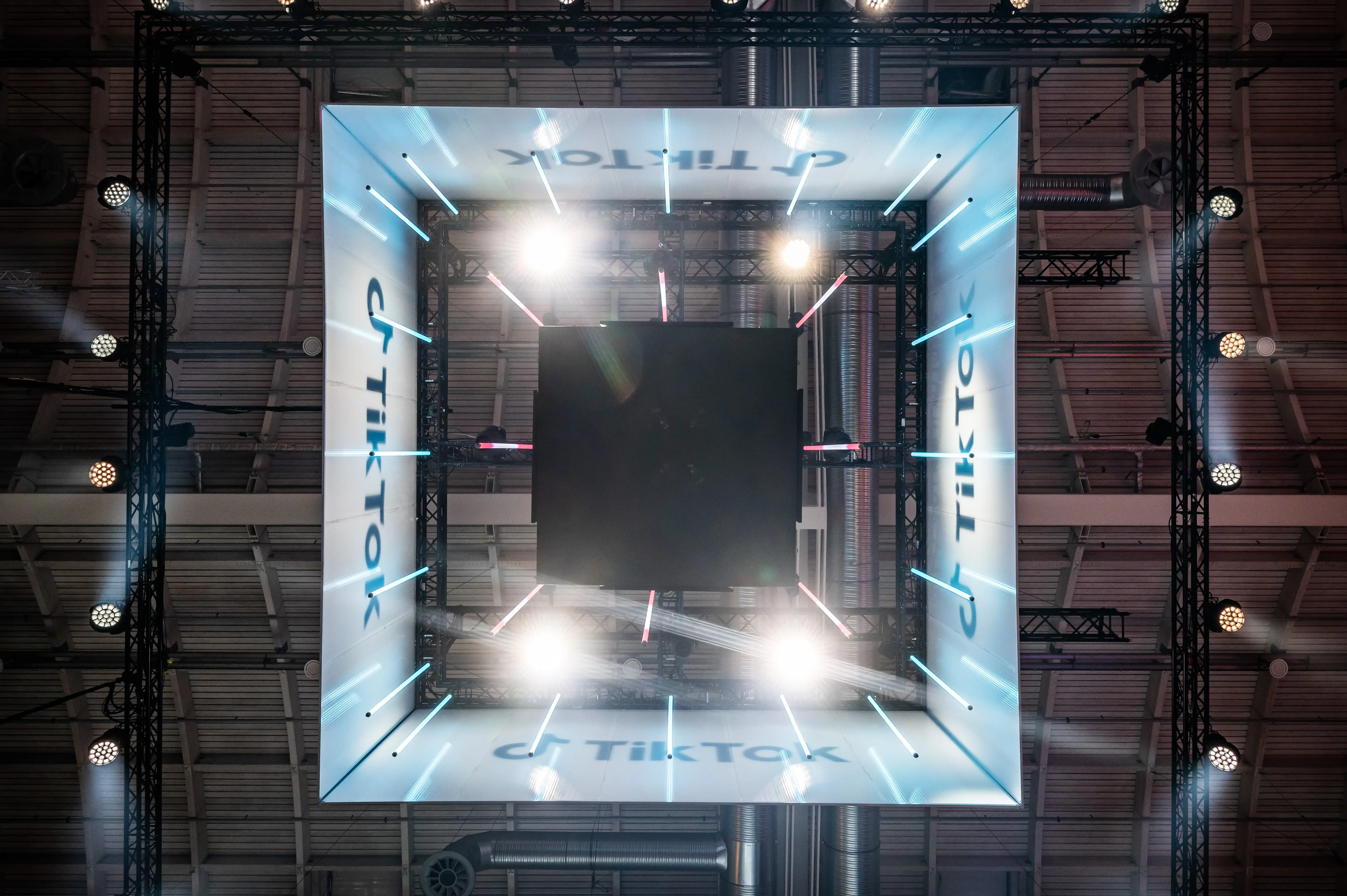 View up to the illuminated TikTok truss cube with screens and spotlights under the hall ceiling.