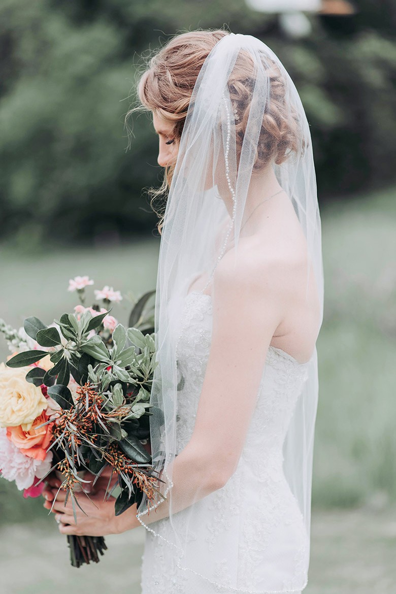 A bride in a white lace wedding dress holds a colorful bouquet of pink, yellow, and orange flowers.