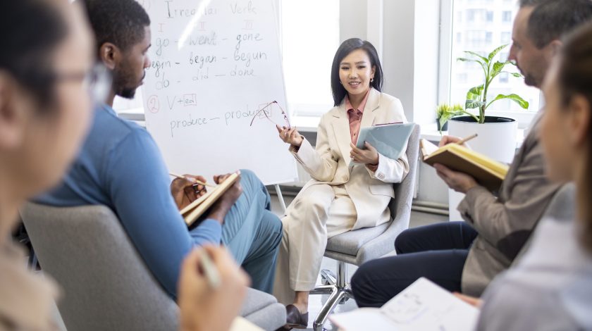 A diverse group of business professionals participates in a brainstorming session, seated in a circle, attentively listening to a presenter in a beige suit, who is explaining strategies with notes on a whiteboard in a modern office setting.