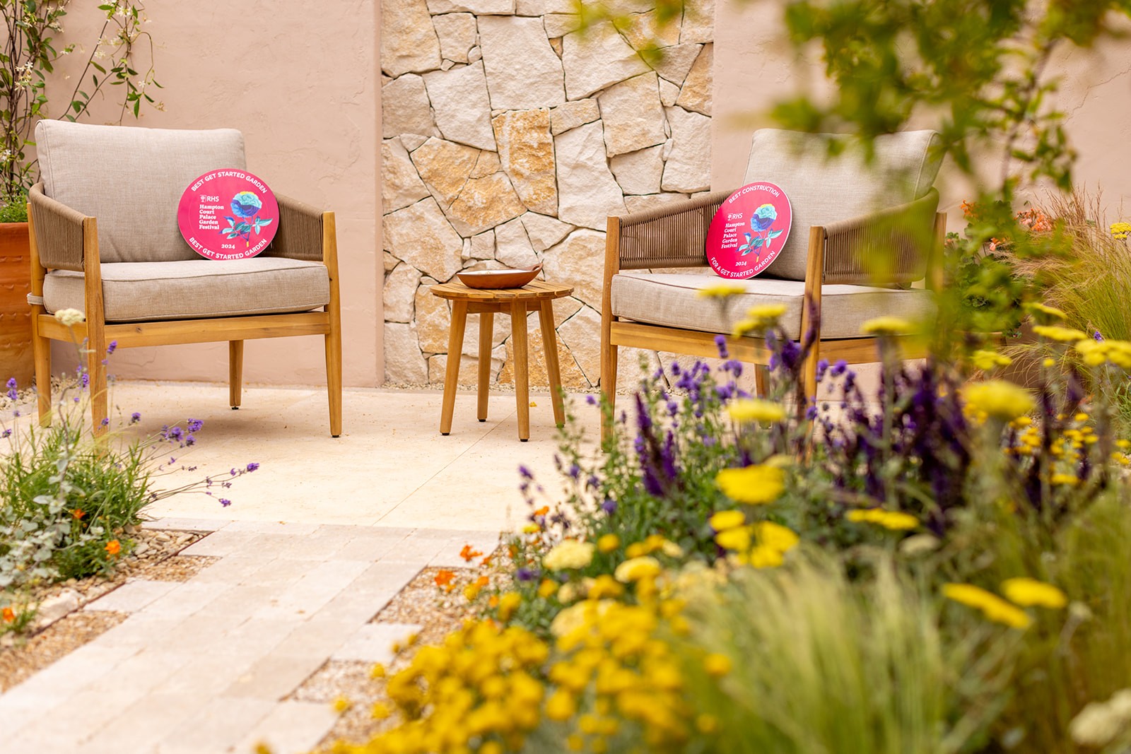 A cozy outdoor space featuring two chairs surrounded by vibrant flowers and greenery, creating a serene atmosphere.