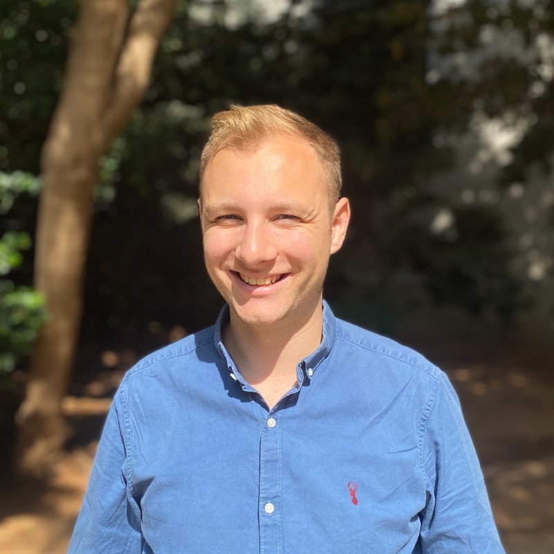 Smiling person in a blue shirt standing outdoors with trees in the background.