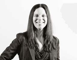 A black and white portrait of a woman with long dark hair, smiling at the camera. She is wearing a dark blazer over a lighter top. The background is plain white.