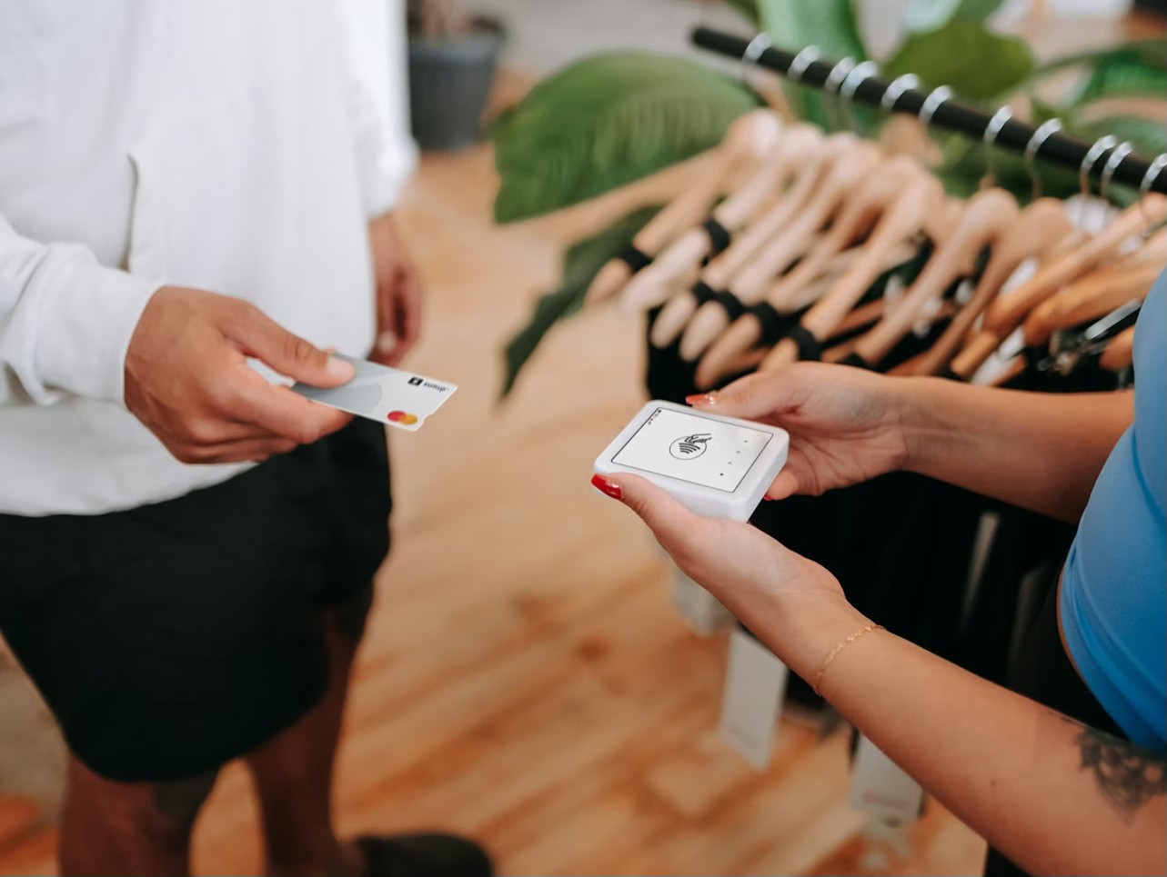 A person holds a smartphone displaying a payment app, while another person presents a credit card, in front of a rack of hanging clothes inside a retail store, illustrating a digital transaction in a shopping environment.