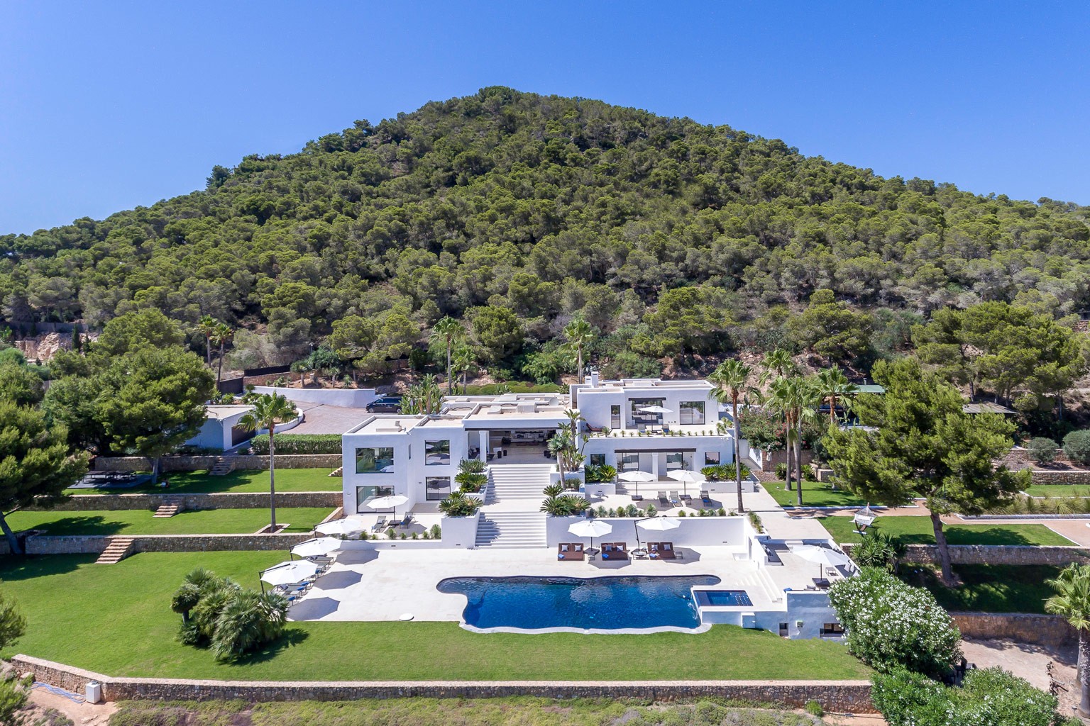 Modern white luxury villa estate with pool and sun terraces set against green hillside landscape near Ibiza Town.
