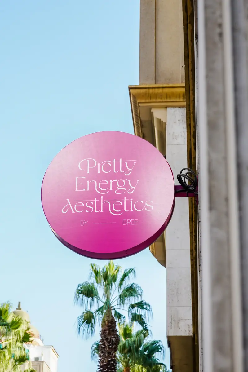 Round pink Pretty Energy Aesthetics shop sign hanging from a building beneath palm trees