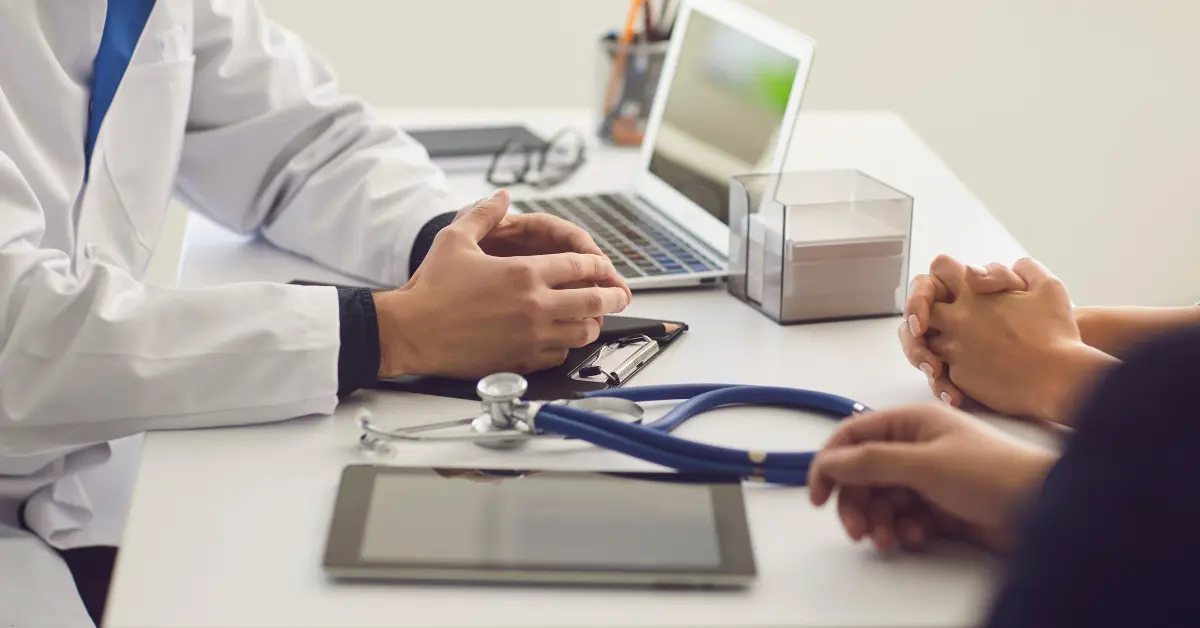 Doctor consulting with a patient at a desk with a stethoscope, clipboard, and laptop.