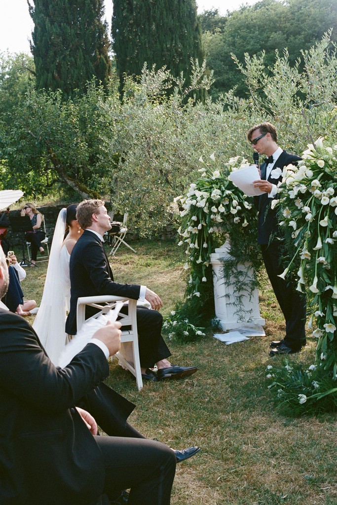 Wedding officiant reading vows in front of couple beneath a floral arch in an olive grove ceremony