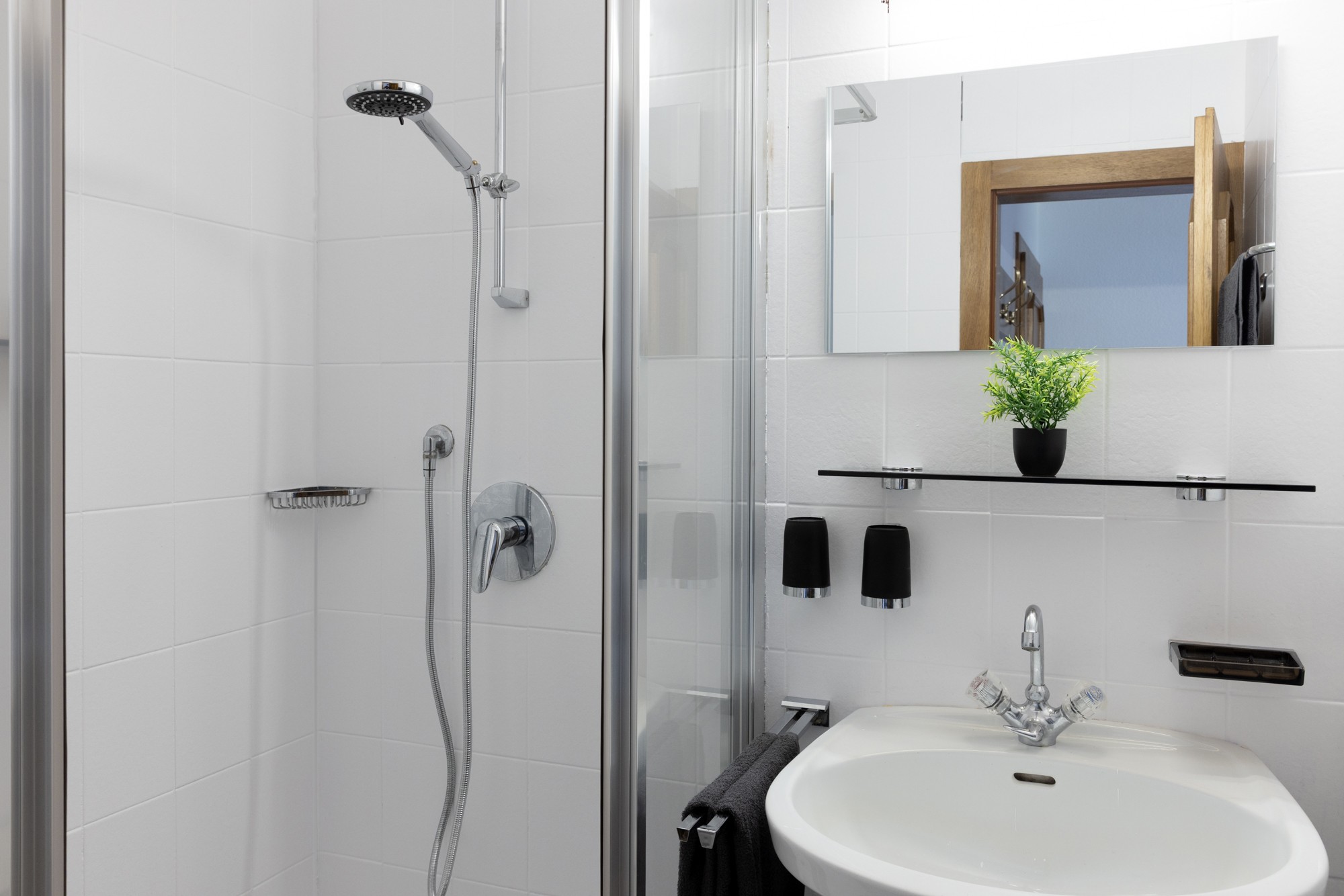 Ein modernes weißes Badezimmer verfügt über eine elegante Glasdusche, ein wandmontiertes Waschbecken mit glänzenden Chromarmaturen, einen rechteckigen Spiegel und eine kleine grüne Pflanze auf einem schwarzen Regal.