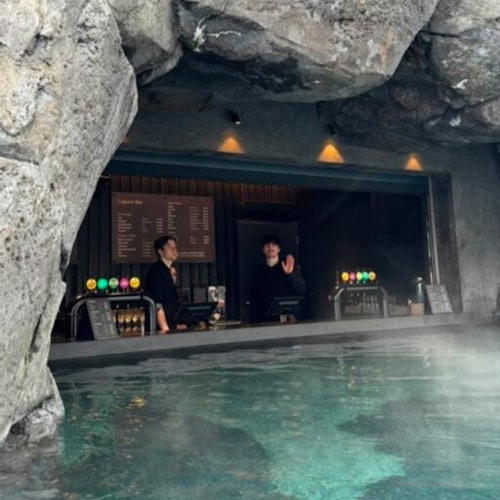 Dos camareros están detrás de una barra ubicada dentro de una cueva rocosa, con una piscina en primer plano y un menú en la pared detrás de ellos.