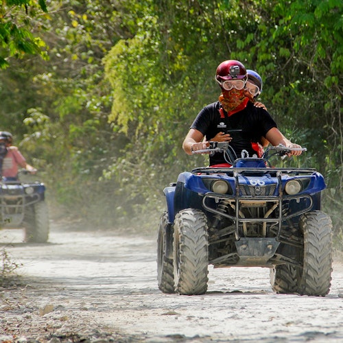 Duas pessoas andando de quadriciclo em um caminho de terra em uma área florestada, com outro quadriciclo e piloto visível ao fundo.