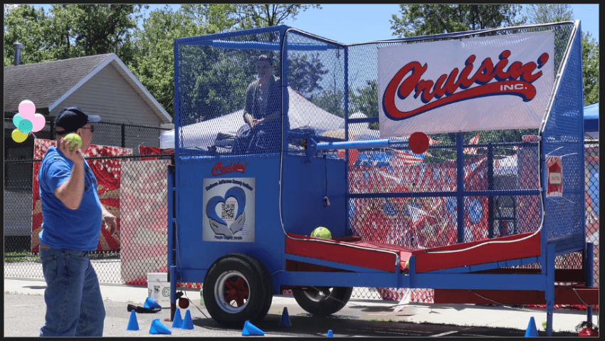 A blue mobile dunk tank trailer setup outdoors with a white banner reading 'Cruisin' Inc.' hung across the safety netting. The side of the tank features waves and Kindness's heart logo.