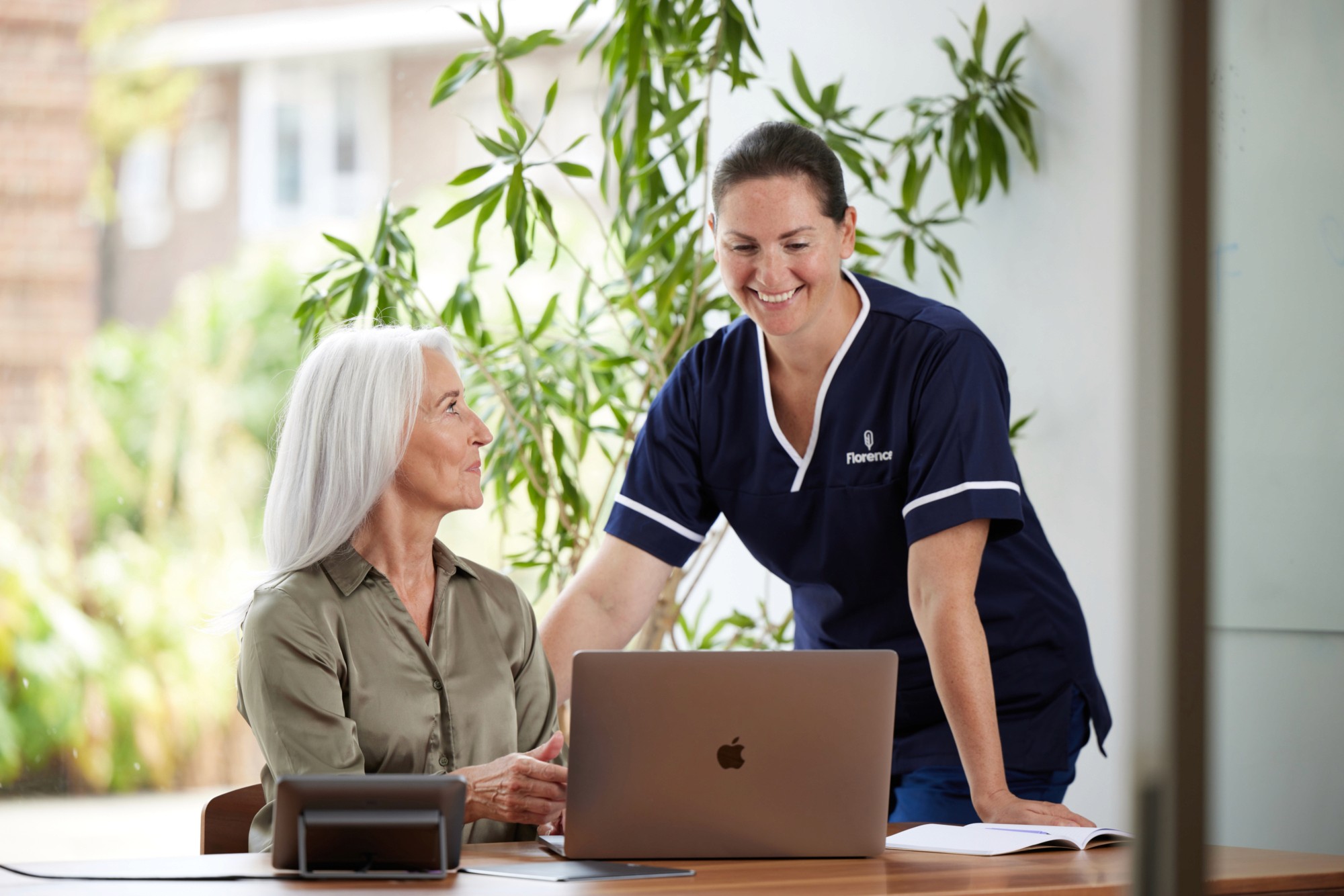 A smiling Florence nurse in navy uniform standing beside an older woman seated at a desk with a laptop, both engaged in a warm interaction