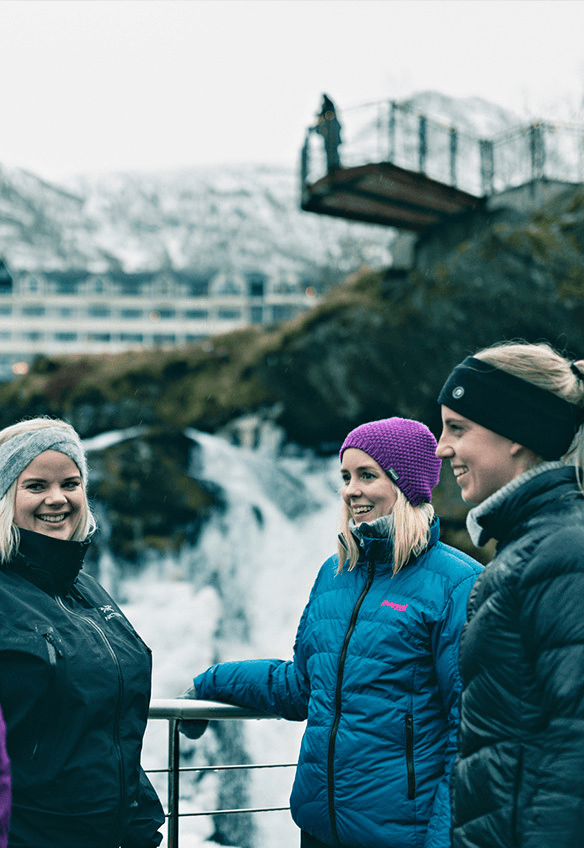 Four people stand together outdoors, smiling and talking, near a waterfall and a bridge in a scenic landscape.