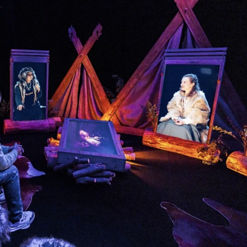 黑暗的房间里，装裱的屏幕展示着身穿毛皮服装的人物，周围布置有原木道具和仿制篝火场景。