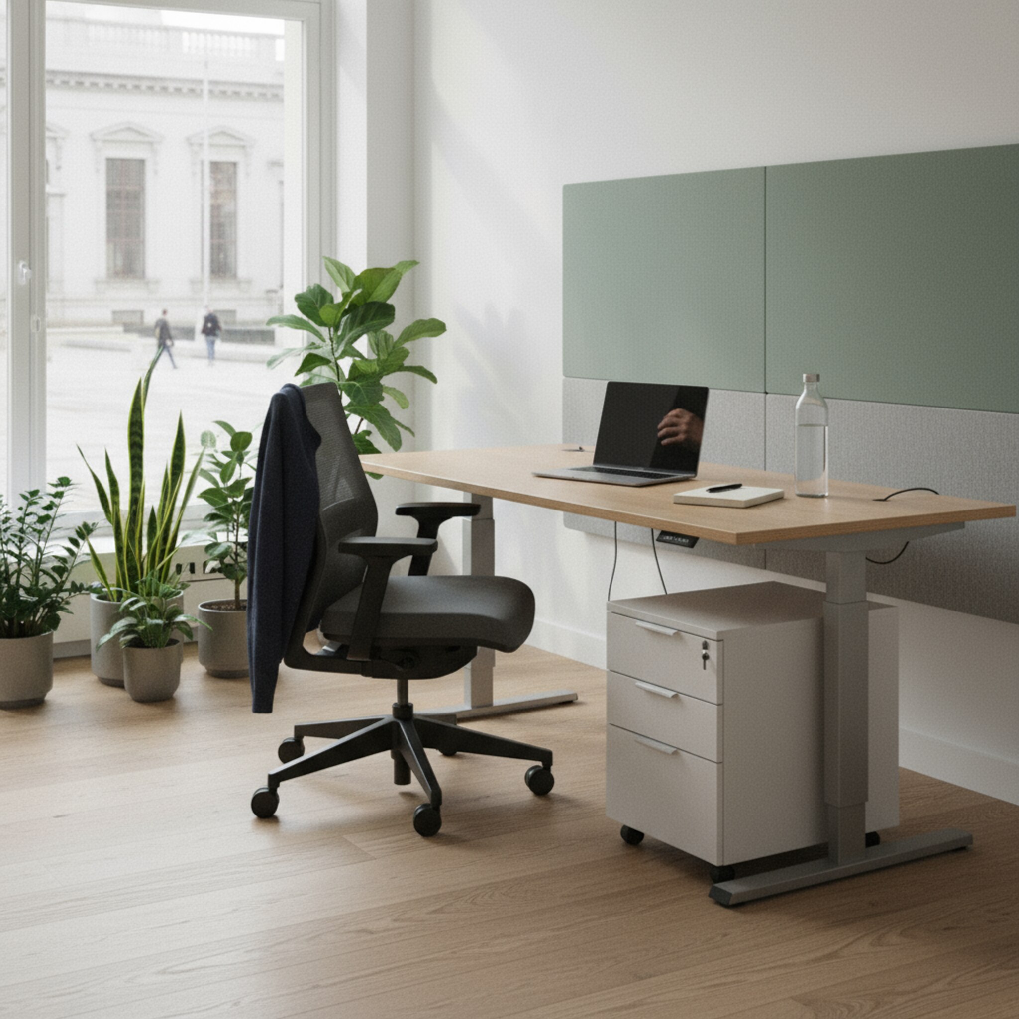 Ein ruhiger Arbeitsbereich im Rathaus zeigt einen höhenverstellbaren Tisch nahe einer großen Fensterfront. Akustikpaneele und eine kleine Begrünung schaffen eine abgeschirmte Zone. Ein ergonomischer Stuhl steht perfekt ausgerichtet, daneben wartet ein leerer Rollcontainer. Das weiche Morgenlicht betont klare Linien und konzentrierte Atmosphäre.