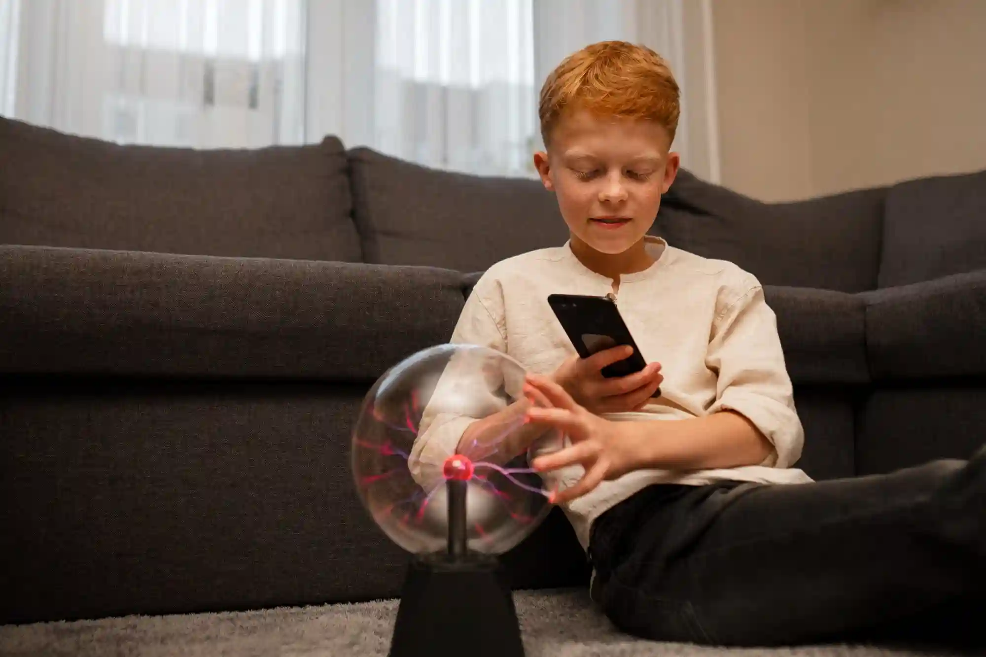 A young boy uses a smartphone while interacting with a plasma globe, illustrating the integration of technology and learning