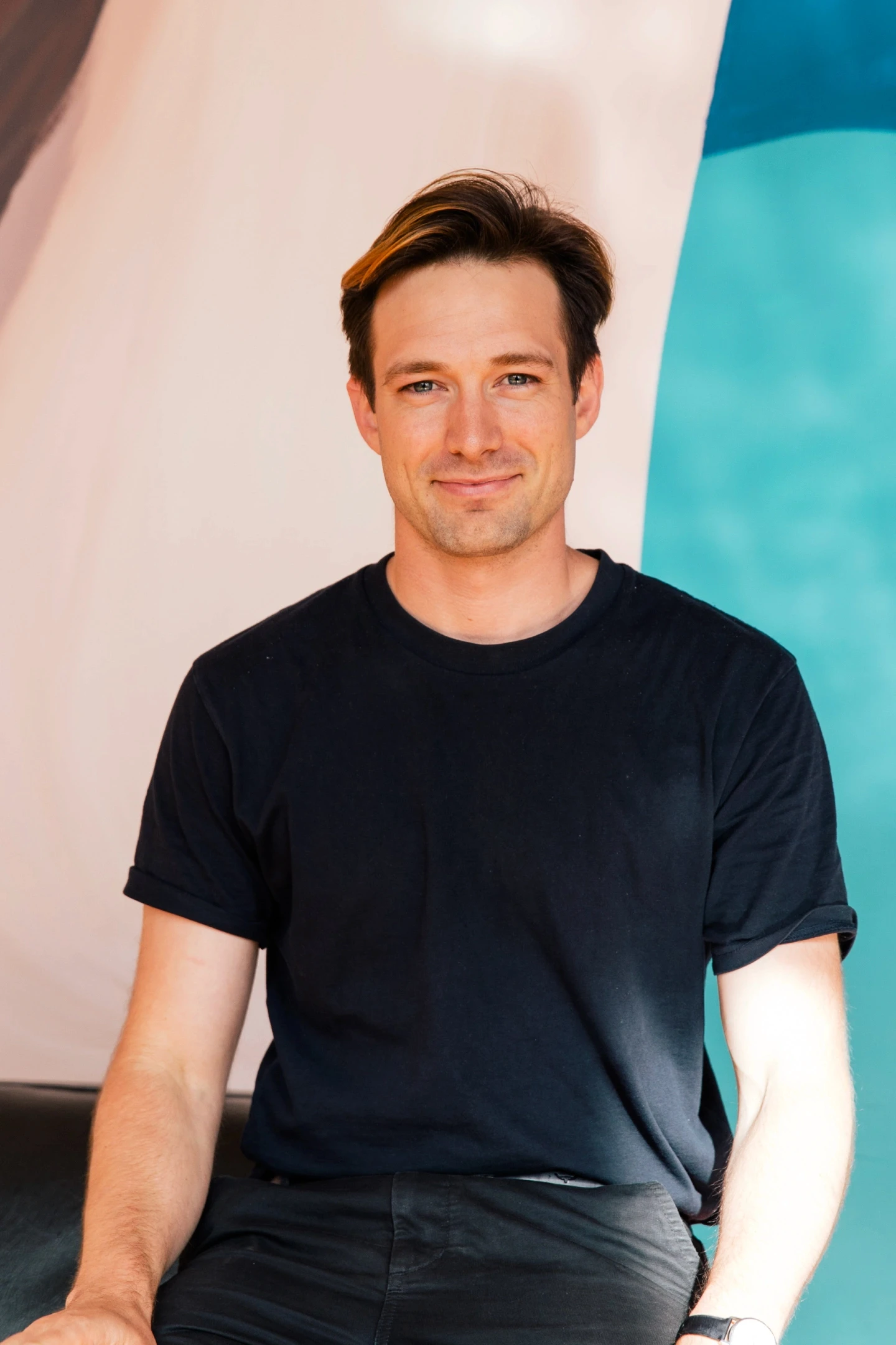 A person wearing a black t-shirt is sitting casually against a colorful background featuring shades of white and teal.