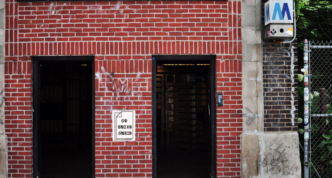 A building made from red bricks with a couple of entrances, a white poster with a note on it, a 3D sign with the letter M and some green leaves near.