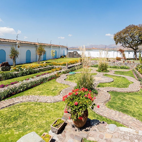 葱鬱的花園有石石徑、五彩繽紛的花朵和盆栽植物，周圍是白色的建築，藍色的門在清澈的天空下。