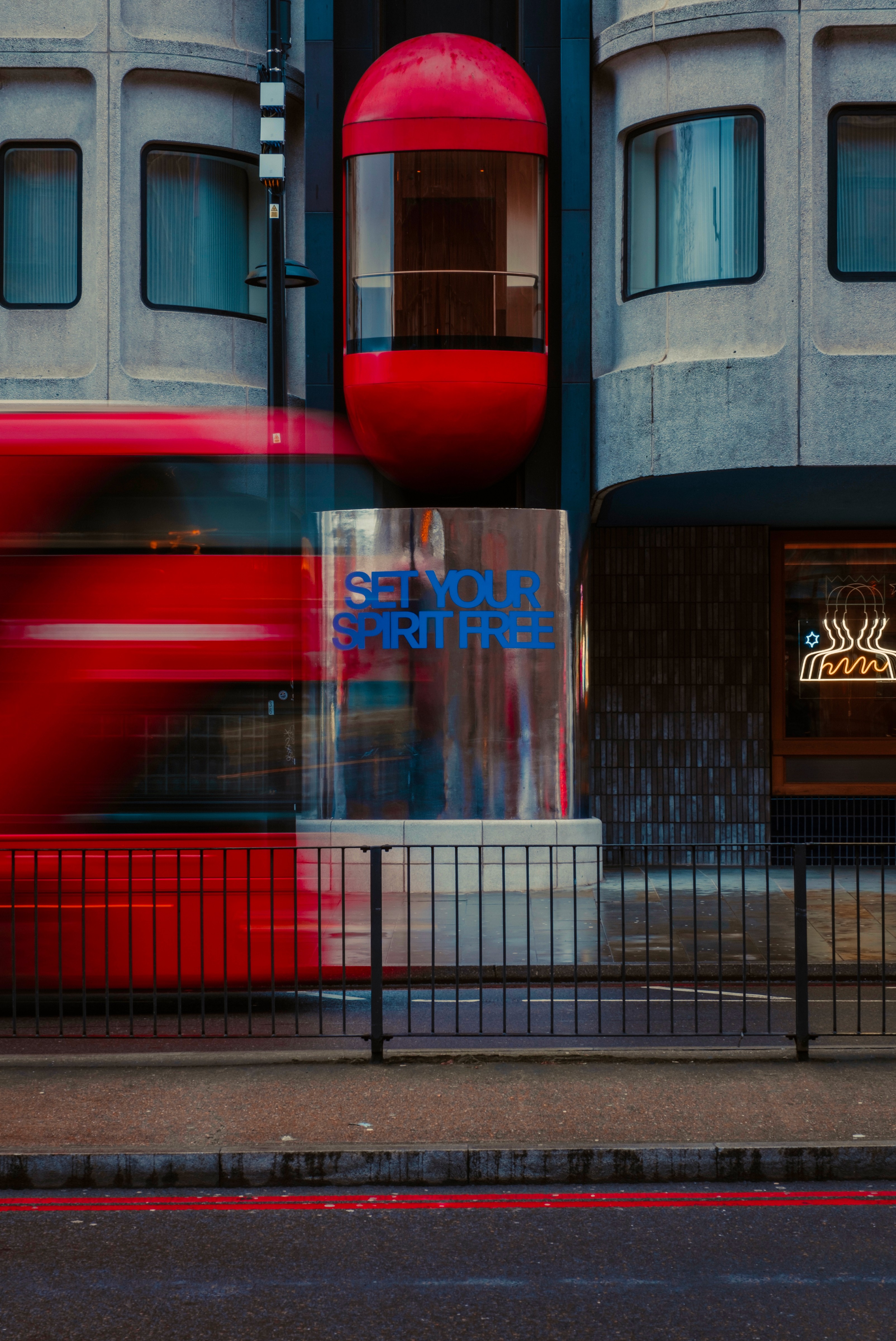 Fotografia uliczna przedstawiająca budynek w Londynie z czerwonym balkonem, rozmazany jadący czerwojy autobus oraz witrynę z napisem "SET YOUR SPIRIT FREE"