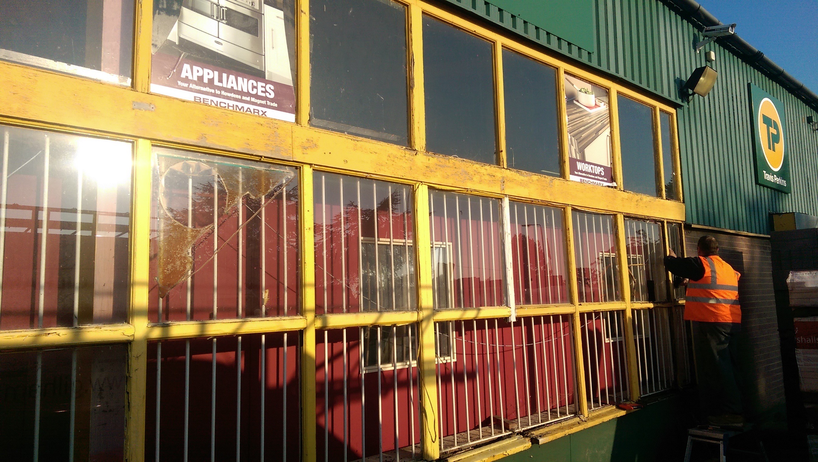 The original shopfront before replacement: a timber and steel framed screen in flaking yellow paint, with cracked and boarded panes and security bars fitted behind the glass
