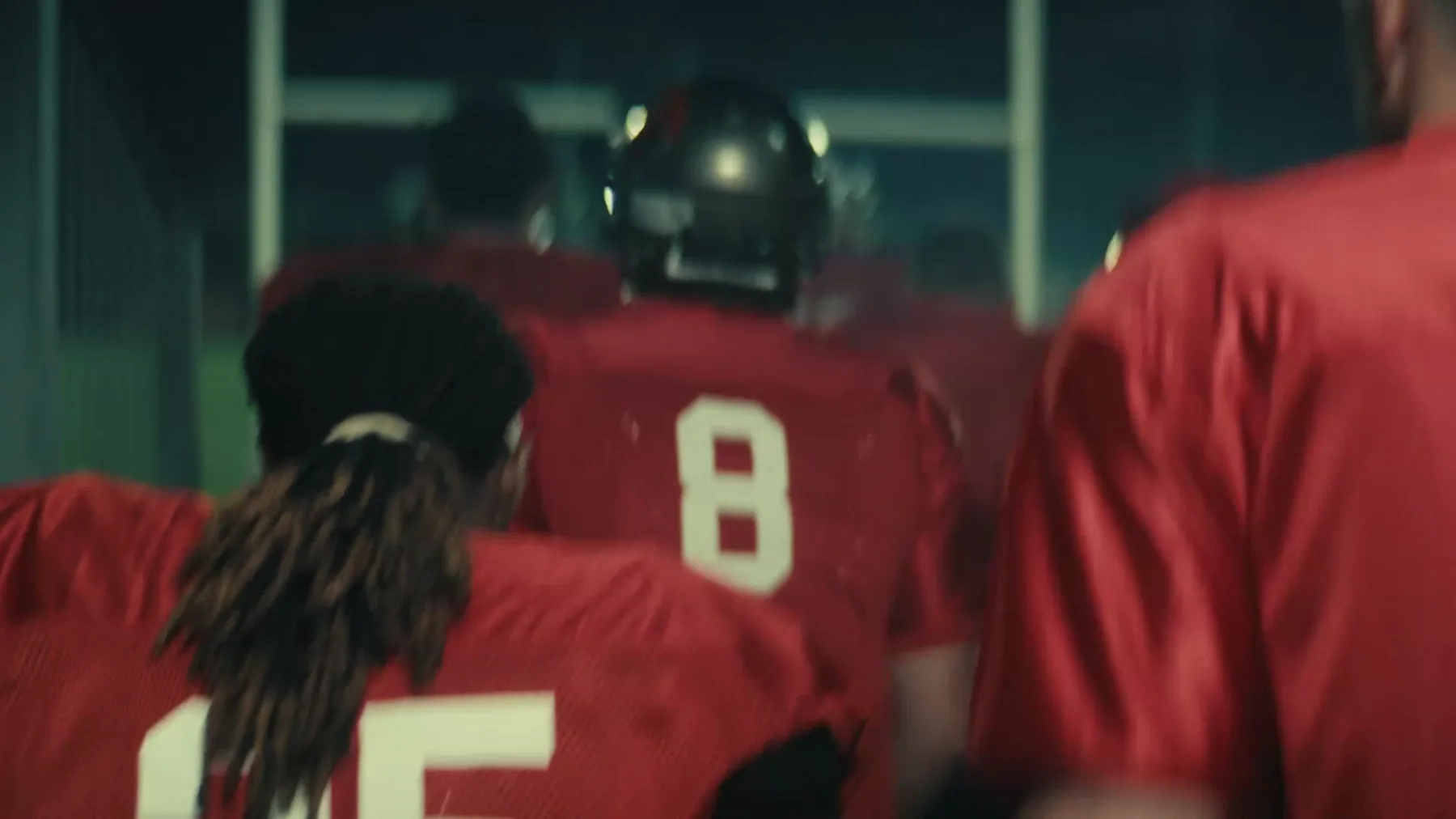 Rear view of football players in red jerseys walking through a tunnel into a stadium, haloed by green smoke and silhouetted against the field lights.