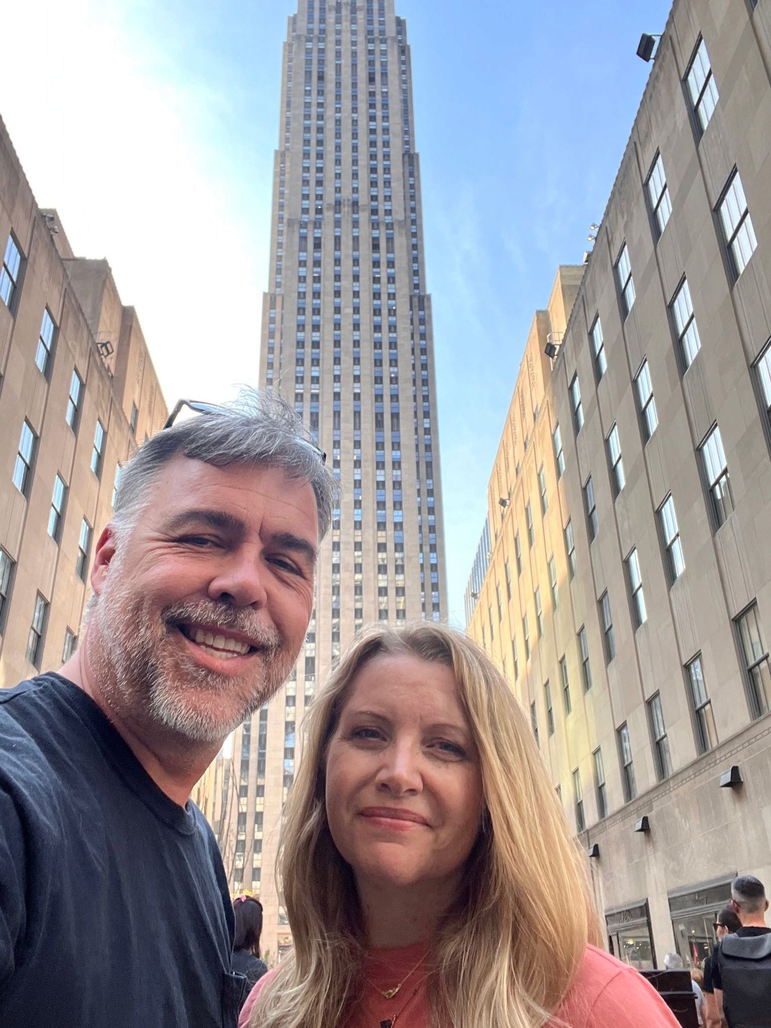 Couple smiling in front of Rockefeller Center skyscraper on sunny city street.