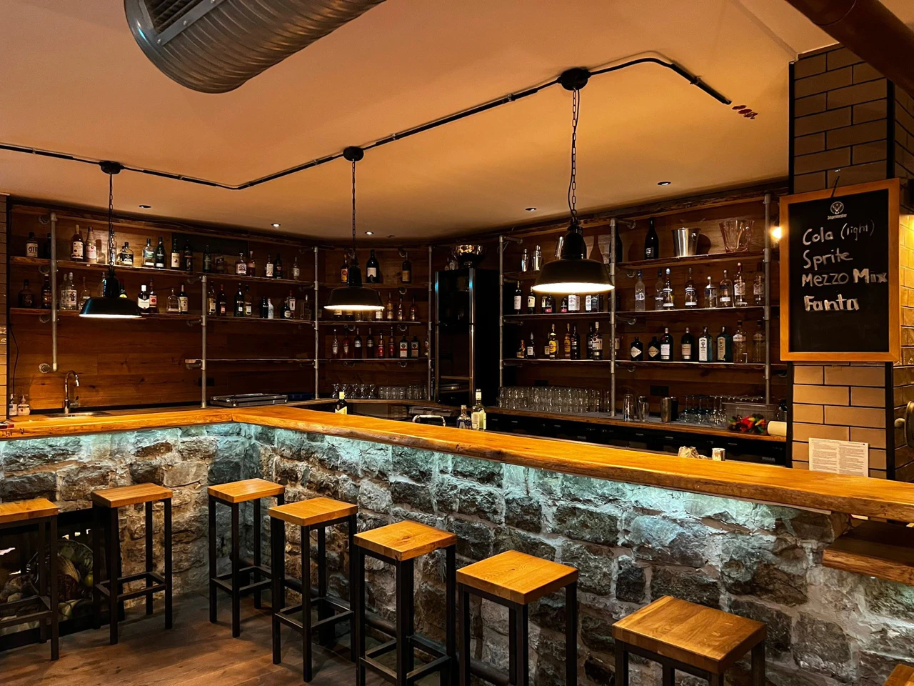 Moderne Bar mit rustikalem Design, Steinmauer-Elementen, Holztheke und Regalen voller Spirituosenflaschen, beleuchtet von hängenden Lampen.