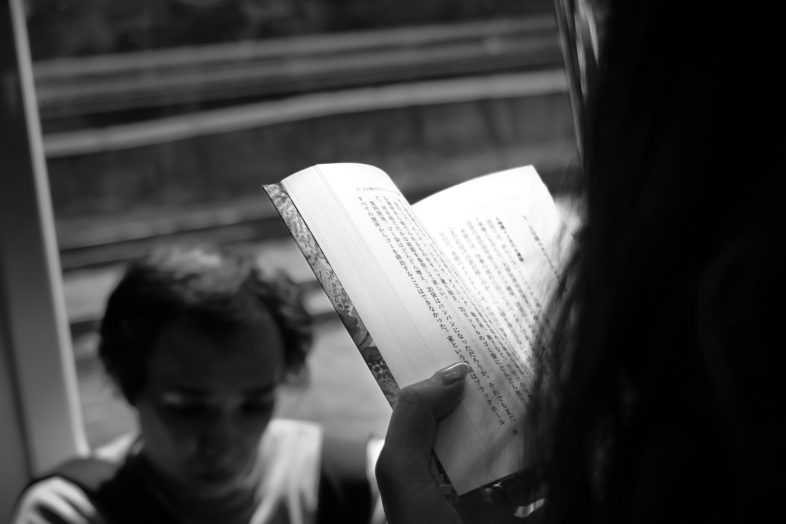 Black and white photograph of a person reading a book inside a train while another figure sits blurred behind them.