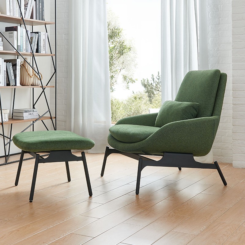 Modern olive green lounge chair with matching ottoman and lumbar pillow, featuring black sculptural legs, set in a bright living room with light wood floors and an industrial bookshelf.