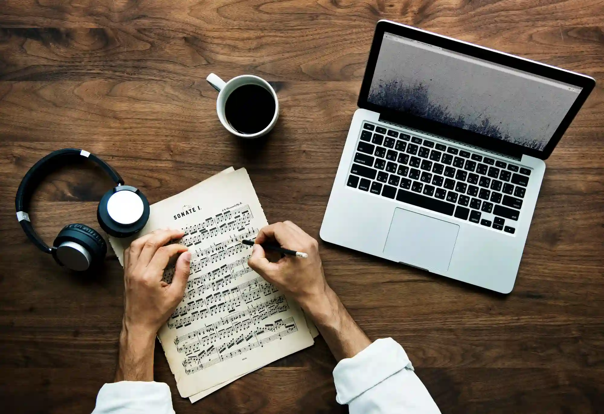 Top-down view of a musician writing sheet music at a wooden desk with a laptop, headphones, and coffee.