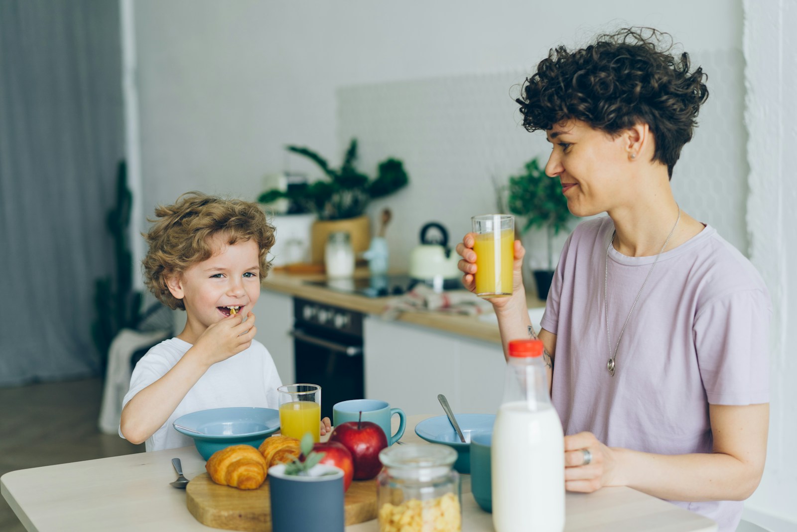 Young child sitting at a bright kitchen table eating breakfast with a parent, with cereal, fruit and juice on the table