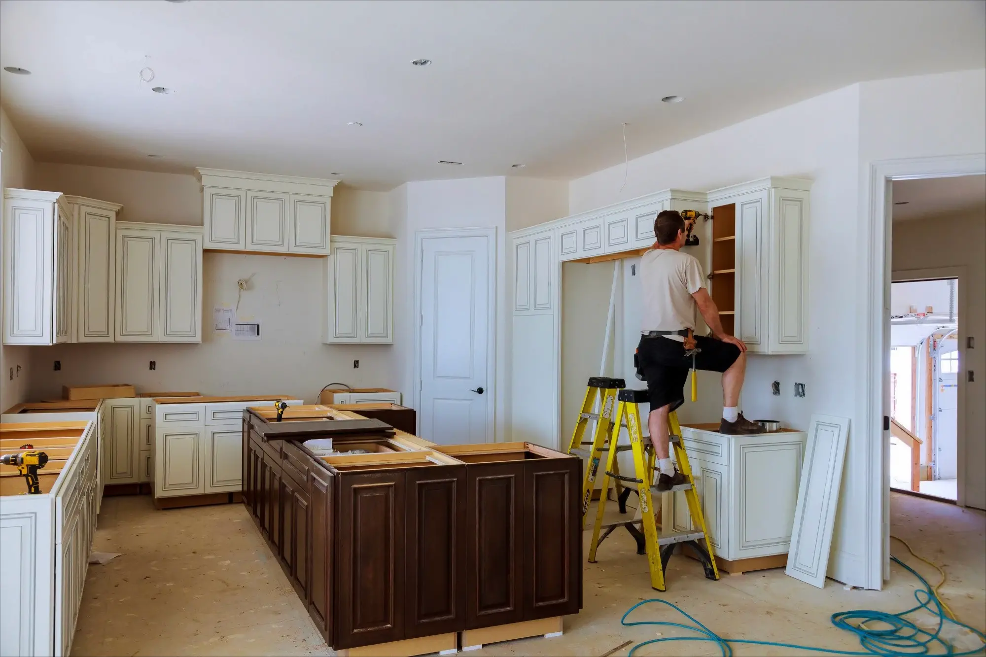 A man installs cabinets in a newly remodeled kitchen, standing on a ladder surrounded by tools and construction materials. The image represents using cash-out refinancing from Chris Lewis Home Loans to fund home renovations and improve property value.