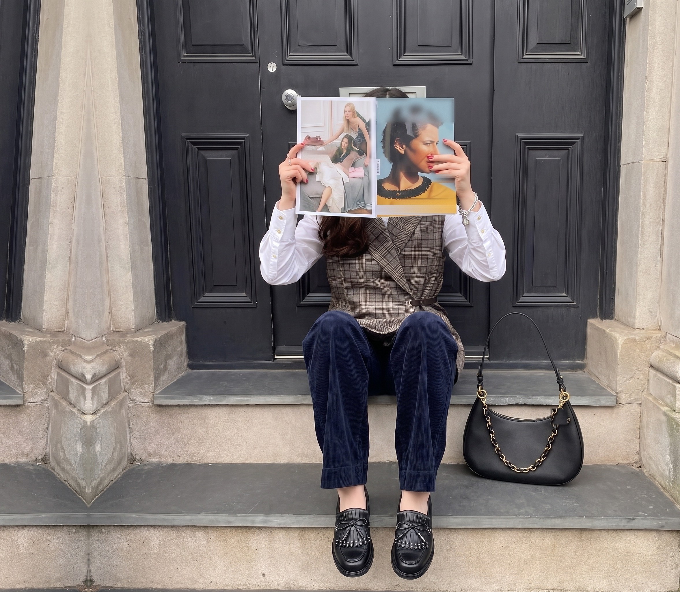 Ragazza seduta sui gradini davanti a un portone nero con gilet a quadri, camicia bianca, pantalone in velluto blu e mocassini, mentre sfoglia una rivista