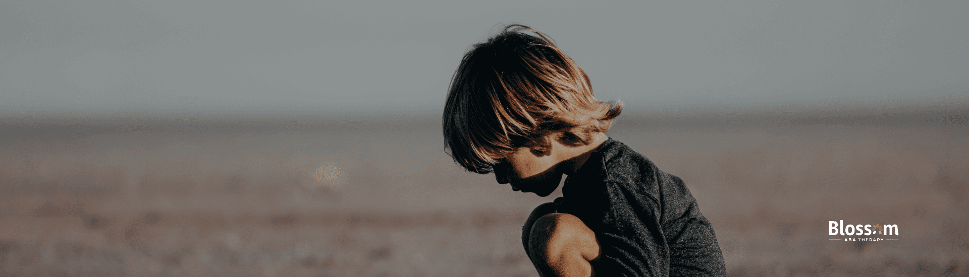 Autistic young boy crouches on sandy beach, focused on the ground in a quiet moment of play after ABA therapy.