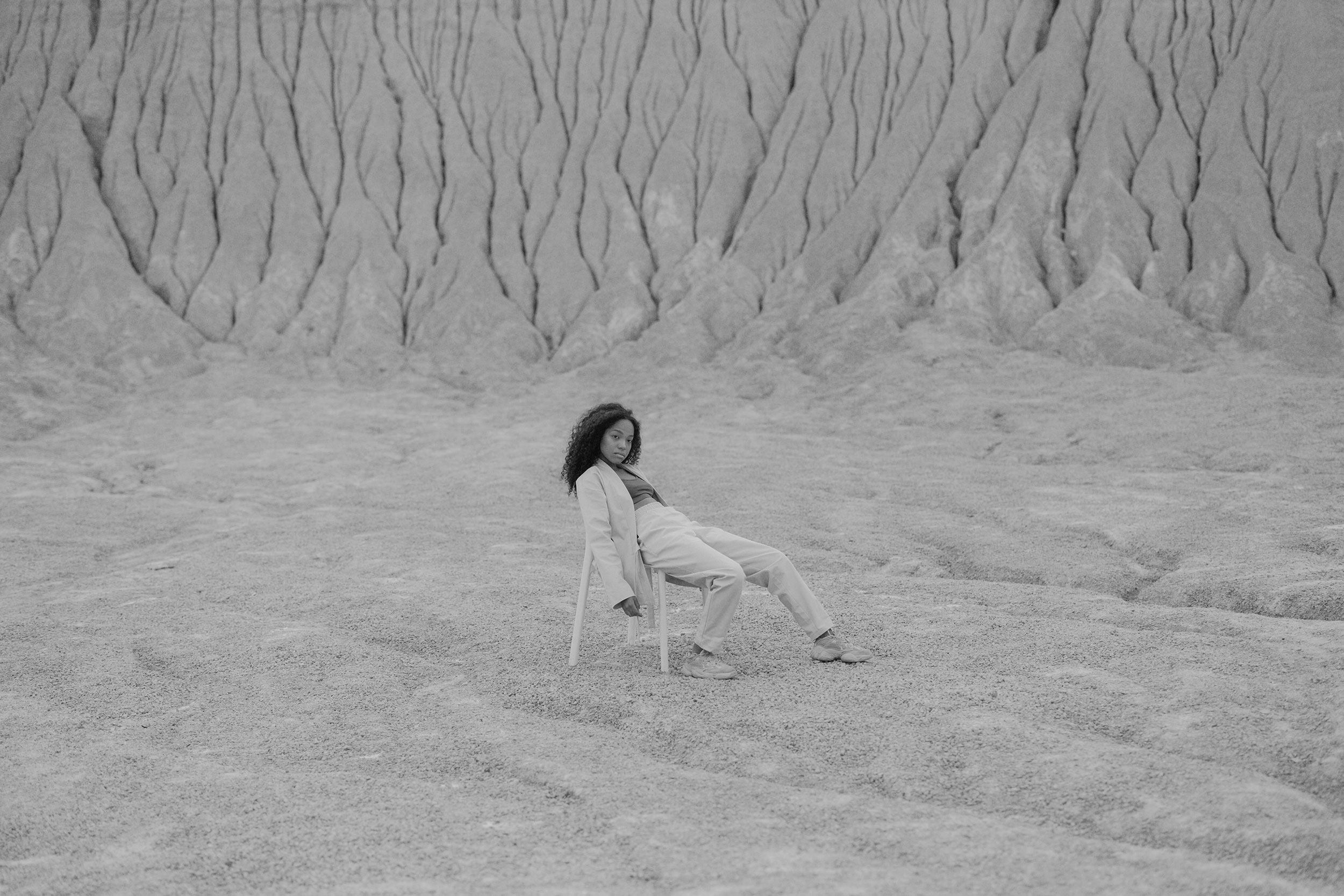 A black and white photo of a person with curly hair sitting on a chair in a barren, textured landscape.