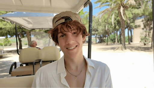 Person smiling at the camera while sitting outdoors, wearing a light-colored shirt and a cap, with palm trees visible in the background.