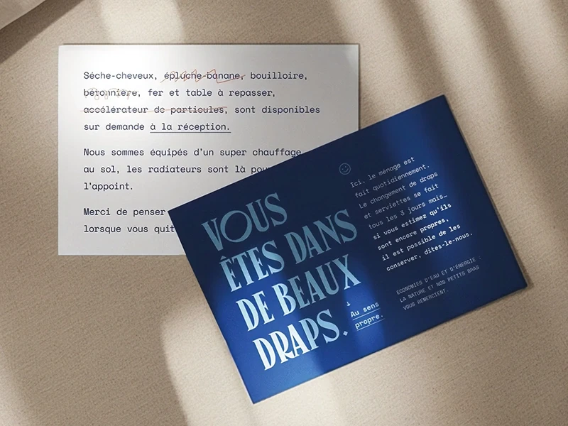 Deux cartes de communication créées pour l’Hôtel Outa : une carte blanche avec une liste de services barrés de façon humoristique et une carte bleue avec le message typographique “Vous êtes dans de beaux draps”, posées sur un drap beige.