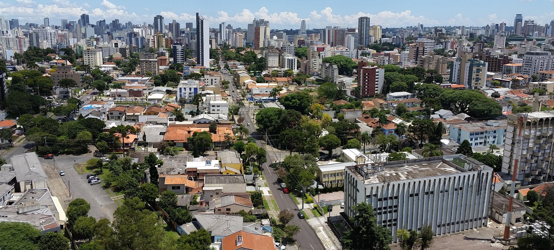 Vista aérea de área urbana com prédios residenciais e comerciais, ruas arborizadas e horizonte da cidade ao fundo.