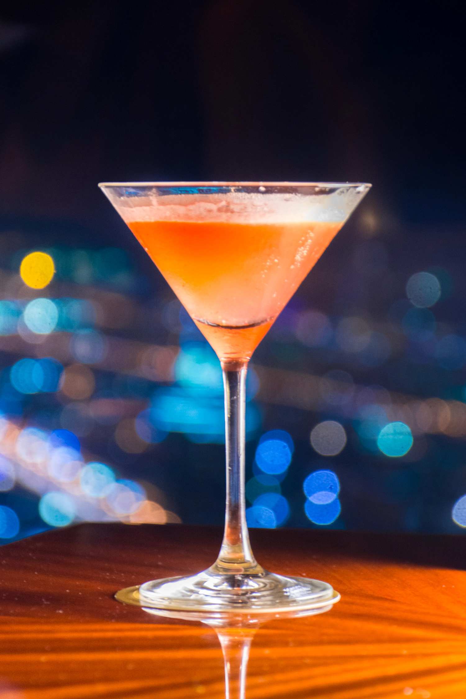 A cocktail in a martini glass sits on a wooden table with a blurred city skyline and bokeh lights in the background.