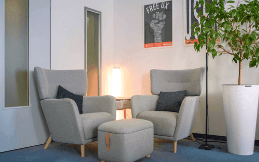 Zwei graue Sessel mit dunklen Kissen und ein grauer Hocker vor einer weißen Wand mit Poster und großem Topfpflanze