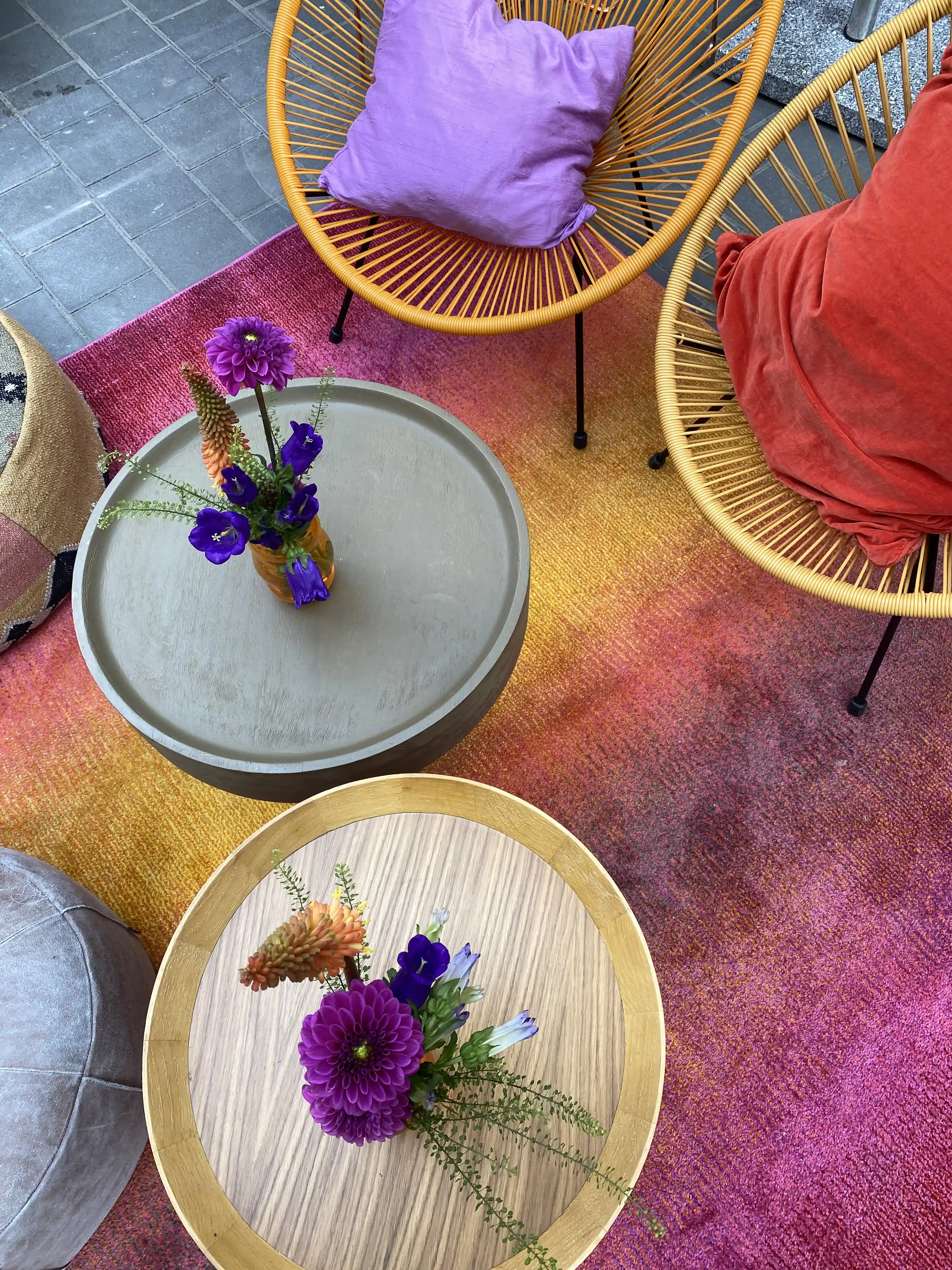 Top view of rattan chairs with colourful cushions and side tables with flower vases on an ombre rug.