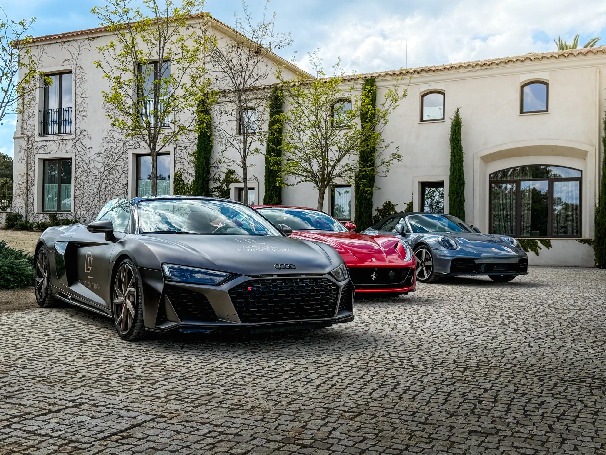 Audi R8 grise, voiture rouge et coupé gris garés devant une grande villa blanche