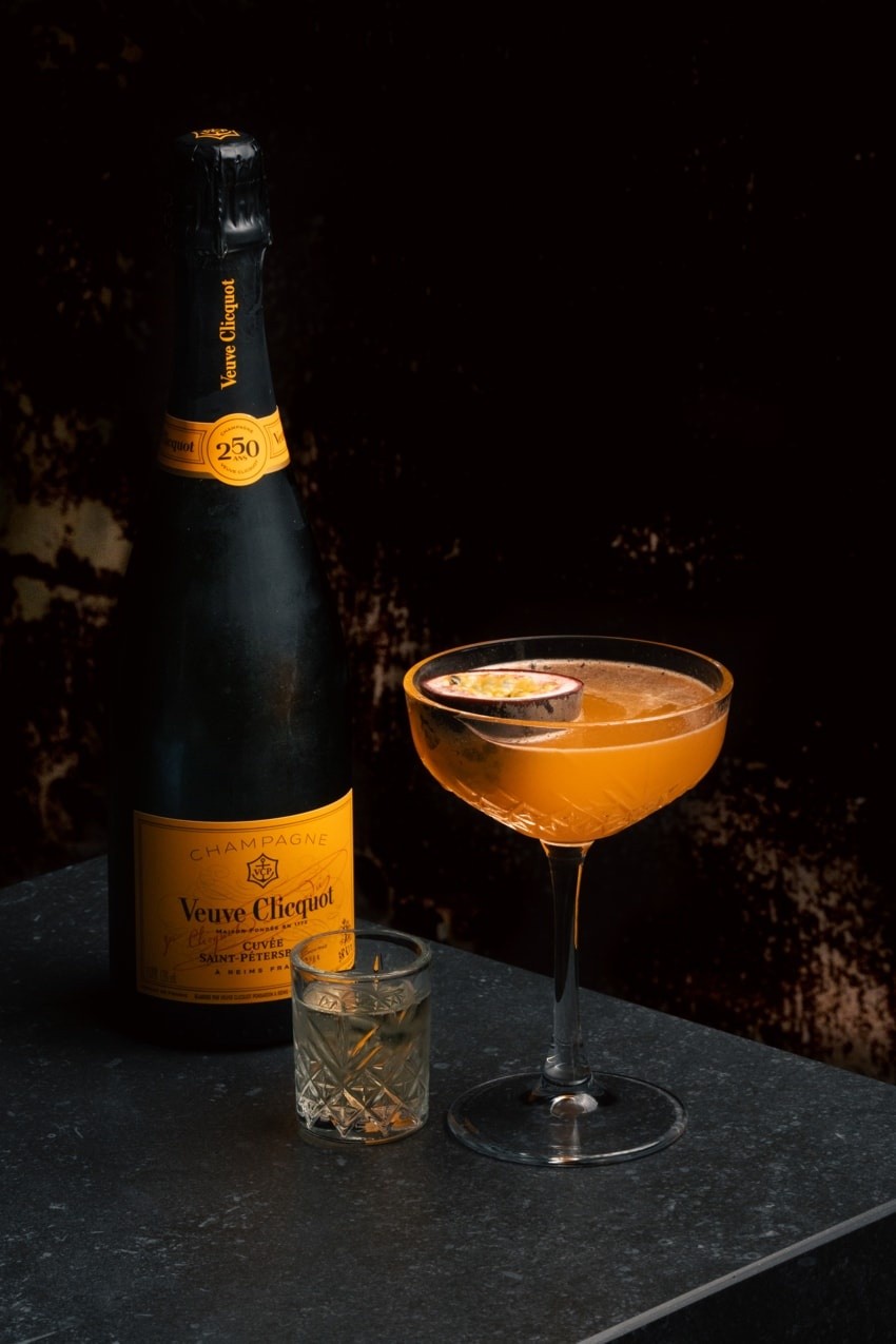 Cocktail arancio servito in calice con guarnizione frutto della passione, accompagnato da shot di Veuve Clicquot Saint Petersbourg