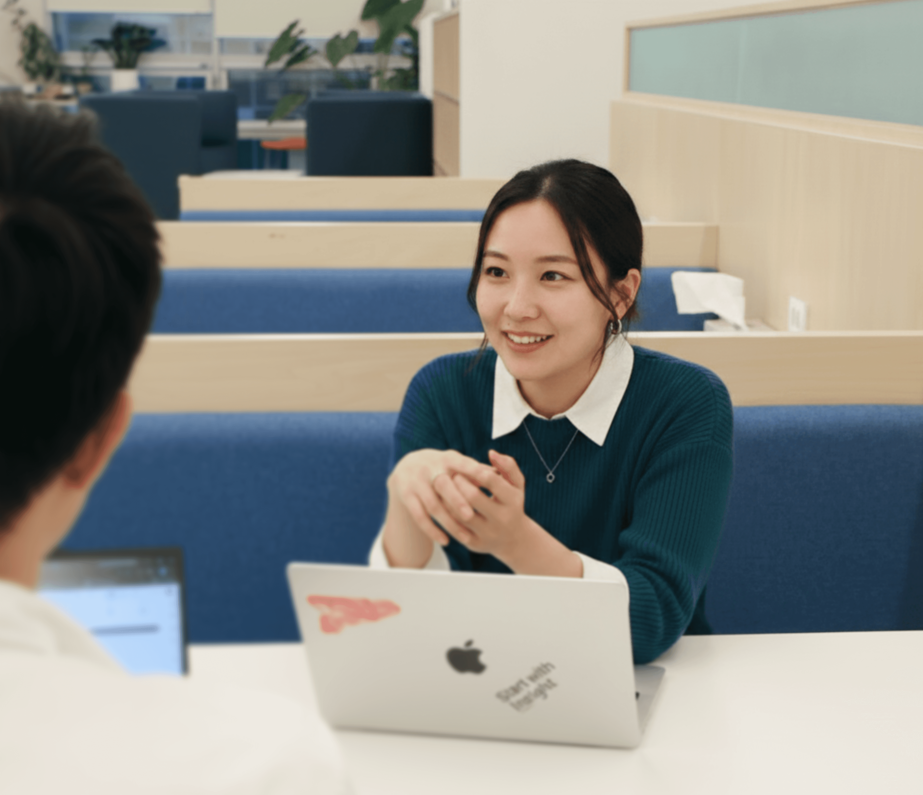 A woman with long hair sits at a table, smiling and engaging in conversation, with a laptop in front of her.