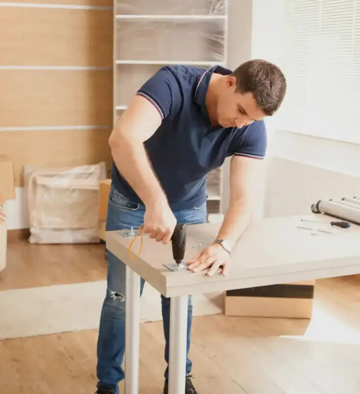 Man using an electric drill to assemble a wooden desk in a bright home office setting.
